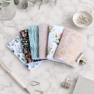 Può includere: Un set di cinque piccoli taccuini disposti a ventaglio, con motivi vari tra cui vita marina, punti blu astratti, foglie disegnate a linee sottili, stampe floreali e rose in lamina d'oro, tenuti insieme da una clip in metallo argentato.