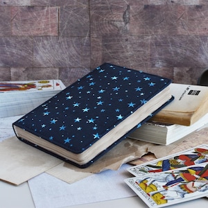 May include: A dark blue book with a star pattern cover, resting on a table with other books and tarot cards. The book has a soft cover with a pattern of blue stars. Other books and cards are scattered around.