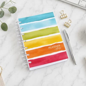 Könnte beinhalten: Ein scheibengebundener Planer mit horizontalem Aquarell-Streifenmuster in Türkis, Hellblau, Limettengrün, Gelb, Orange und Magenta. Auf dem Einband steht der weiße Text PLANNER 365. Der Planer ist mit durchsichtigen Kunststoffscheiben gebunden.