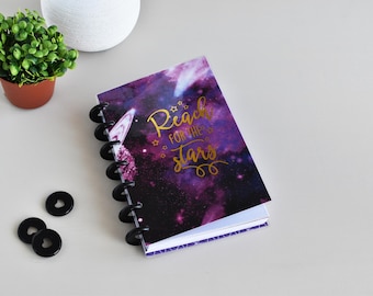May include: A spiral-bound notebook with a galaxy-themed cover featuring the text "Reach for the stars" in gold lettering. The notebook is open, revealing white pages. Black disc-shaped rings and extra discs are visible.
