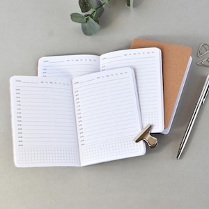 Peut inclure: Deux agendas ouverts avec des pages blanches présentant des horaires de 07:00 à 18:00, les jours de la semaine et une section quadrillée en bas. Un carnet marron fermé, un stylo en métal argenté et deux pinces argentées sont disposés à côté.