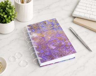 May include: A disc-bound notebook featuring a purple watercolor background with a gold foil floral and text overlay. The cover is secured with clear plastic discs along the left side, showcasing a vibrant design with visible script and botanical line art.