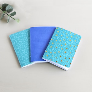 Könnte beinhalten: Drei kleine Notizbücher, die fächerförmig angeordnet sind. Das linke Notizbuch ist hellblau mit einem tonalen Blumenmuster. Das mittlere Notizbuch ist einfarbig mittelblau mit einem dunkelblauen Gitter. Das rechte Notizbuch ist hellblau mit gelbem Gänseblümchenmuster.
