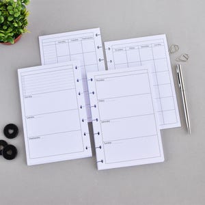 Puede incluir: Insertos de planificador blancos con un diseño semanal, con secciones para cada día y una lista de tareas. Un bolígrafo plateado y clips están junto a las páginas del planificador. Anillos negros encuadernados en disco y una pequeña planta en maceta también están en la imagen.