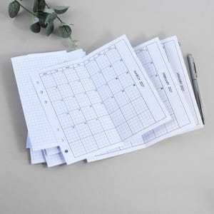 Könnte beinhalten: Ein Stapel weißer Planer-Einlagen aus Papier mit Gittermuster und Lochungen an der linken Seite. Die Seiten zeigen Monatskalender für Januar, Februar und März 2027. Ein silberner Metallkugelschreiber liegt rechts neben dem Papierstapel.