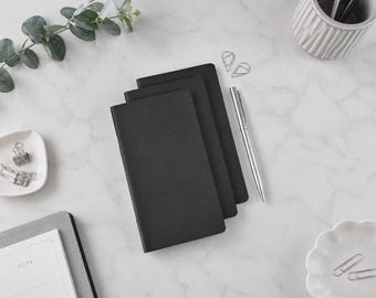 May include: A stack of three black rectangular notebooks with a silver pen placed beside them. The notebooks are plain and solid black. Nearby are silver paper clips, a small white dish, and a gray notepad with the word NOTE printed.