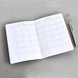 Può includere: Un'agenda aperta che mostra un calendario mensile per gennaio 2026 con una disposizione a griglia. Le pagine presentano testo e linee stampate in nero. Una penna in metallo argentato e due graffette in metallo argentato sono posizionate a destra.