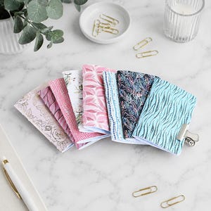 May include: A collection of seven small notebooks arranged in a fan shape, featuring various patterns including floral, geometric, and abstract designs in shades of pink, blue, and black, secured together with a silver metal binder clip.