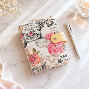 Puede incluir: Un cuaderno rectangular con una cubierta de tela color crema que presenta un estampado de estilo vintage con rosas rosadas, texto en caligrafía negra y sellos postales. El cuaderno incluye una correa amarilla pálida con cierre de metal y un bolígrafo.