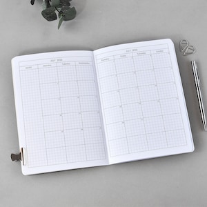 Könnte beinhalten: Ein offenes Notizbuch mit einem Kalenderlayout für Juli 2026. Das weiße Notizbuch hat ein Rastermuster und wird von einem silbernen Stift und Büroklammern begleitet. Ein kleiner Metallclip hält das Notizbuch offen.