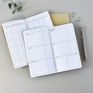 Könnte beinhalten: Offene Notizbücher mit leeren Kalender- und Wochenplanerseiten. Die Notizbücher haben einen beigefarbenen Einband und werden mit silbernen Büroklammern offen gehalten. Ein silberner Stift und zwei silberne Büroklammern liegen ebenfalls auf der grauen Oberfläche.