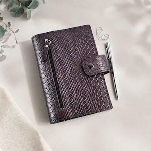 May include: A dark purple notebook with a textured snakeskin pattern, featuring a vertical zippered pocket on the front cover, a snap-button closure strap, and a silver-toned metal pen resting alongside two silver paper clips.