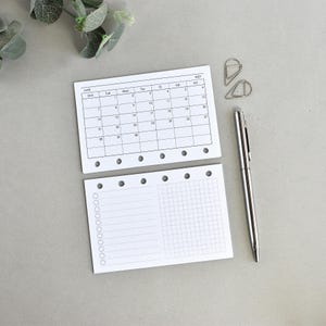 May include: Two white planner inserts with six punched holes along the edge. The top sheet shows a June 2027 calendar grid. The bottom sheet features a checklist with circular bullets on the left and a grid pattern on the right. A silver pen is included.