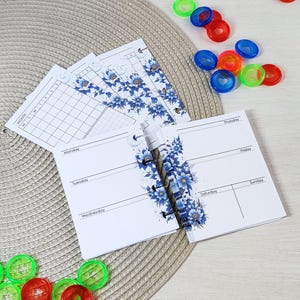 May include: White planner pages with blue floral accents and a monthly calendar layout. The pages are held together with plastic rings. Colorful plastic rings in red, green, and blue are scattered around the planner.