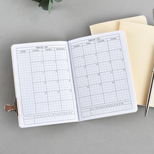 Könnte beinhalten: Offenes Notizbuch mit Kalenderlayout für Februar 2024, offen gehalten mit einer silbernen Klammer. Ein silberner Stift und zwei Büroklammern liegen neben einem geschlossenen, hellfarbigen Notizbuch. Der Hintergrund ist einfarbig grau.