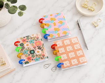 May include: Three small disc-bound notebooks with colorful patterned covers. One features a pink, blue, and yellow abstract mosaic, one has a floral print on a peach background, and one displays a grid of white floral squares on a peach background.