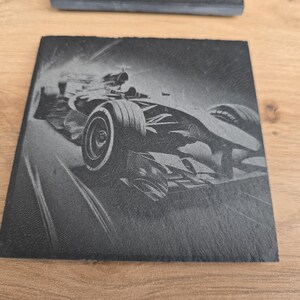 Könnte beinhalten: Ein Schwarzweißbild eines Rennwagens, der auf einer Rennstrecke rast. Das Auto ist in Bewegung, wobei der vordere Reifen im Fokus steht. Das Bild ist auf einen schwarzen Schieferuntersetzer graviert.