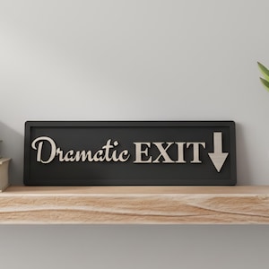 May include: A rectangular black sign with raised, light-colored lettering that reads "Dramatic EXIT" followed by a downward-pointing arrow. The sign features a clean, bold border and is designed for wall mounting or display on a flat surface.