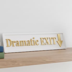 May include: A rectangular white sign featuring the gold-coloured raised text "Dramatic EXIT" followed by a downward-pointing arrow. The sign has a clean, framed border and is positioned horizontally, displaying clear, bold lettering in a serif font.