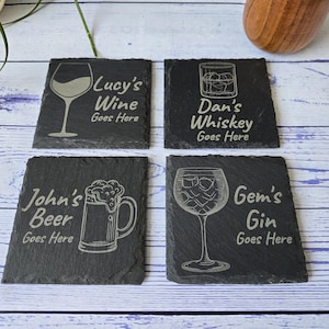 May include: Four black slate coasters with engraved designs and text. Each coaster features a different drink: wine, whiskey, beer, and gin. The text on each coaster reads "Lucy's Wine Goes Here", "Dan's Whiskey Goes Here", "John's Beer Goes Here", and "Gem's Gin Goes Here".
