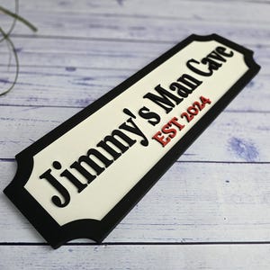 Puede incluir: Un letrero rectangular con borde negro y centro crema. El letrero dice "Jimmy's Man Cave EST 2024" en letras negras y rojas. El letrero está en ángulo, mostrando su diseño dimensional.