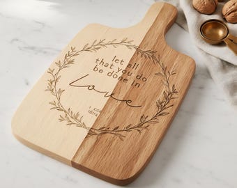 Puede incluir: Tabla de cortar de madera de dos tonos con asa. Un lado es de color claro, el otro es más oscuro. La tabla presenta un diseño de corona floral grabada con las palabras "let all that you do be done in love" y la referencia "1 COR 16:14".