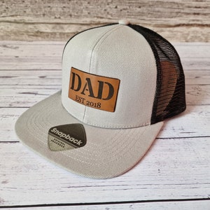 May include: A grey and black trucker hat with a brown leather patch that says "DAD EST 2018".