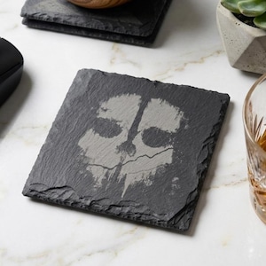 May include: A square, dark grey slate drinks coaster featuring a skull design. The coaster sits on a white marble surface, alongside a black computer mouse and a glass of amber liquid. A succulent plant in a concrete pot is also visible.