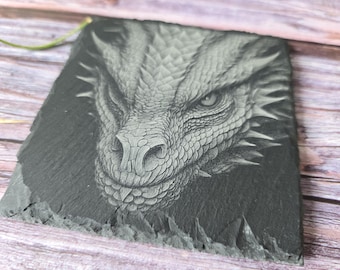 Puede incluir: Un posavasos cuadrado de pizarra gris oscuro con un rostro de dragón grabado. Las escamas, el ojo y el hocico del dragón están intrincadamente tallados, creando un efecto texturizado y tridimensional. Una pieza de decoración única.