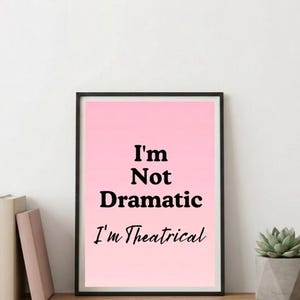 Op de afbeelding: Een ingelijste wandkunstprint met een roze gradiëntachtergrond en de zwarte tekst "I'm Not Dramatic" in een vetgedrukt schreeflettertype en "I'm Theatrical" in een zwart cursief lettertype, gevat in een dunne zwarte rechthoekige lijst.