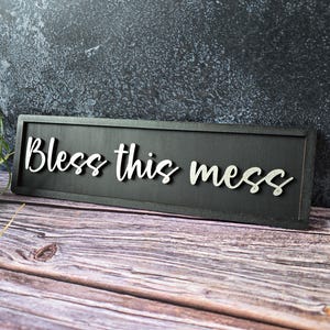 Puede incluir: Un letrero rectangular de madera negra con la frase "Bless this mess" en escritura blanca. El letrero está sobre un fondo texturizado gris y negro, apoyado sobre una superficie de madera.