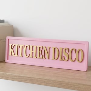 May include: A rectangular pink sign with raised gold lettering that reads KITCHEN DISCO. The sign features a smooth, matte finish on the pink frame and a metallic gold sheen on the letters, presenting a clean, bold, and high-contrast appearance.