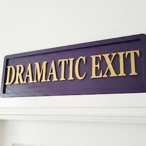 Puede incluir: Un letrero rectangular de madera con las palabras "DRAMATIC EXIT" en letras doradas. El letrero tiene un fondo morado oscuro y está montado sobre una superficie blanca.