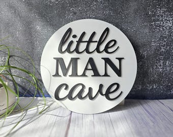 May include: A white, round wooden sign with the words "little MAN cave" in black, stylized lettering. The sign is displayed on a light wood surface with a gray and white textured background. A small potted plant is visible on the left.