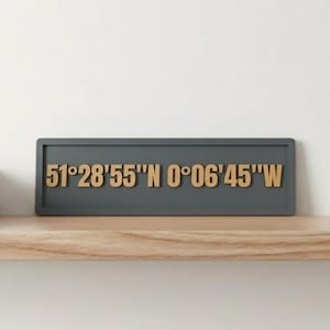May include: A grey rectangular sign displaying the coordinates 51°28'55"N 0°06'45"W in a wooden font. The sign is on a wooden shelf, with books and a plant in the background. The shelf is likely made of wood.