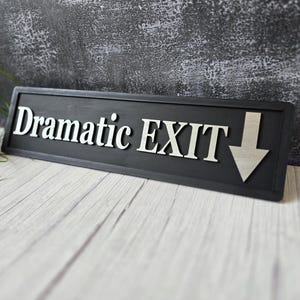 Puede incluir: Un letrero rectangular negro con el texto plateado "Dramatic EXIT" en una fuente con serifa, acompañado de una flecha plateada que apunta hacia abajo en el lado derecho. El letrero tiene un borde elevado y un acabado mate en las letras.