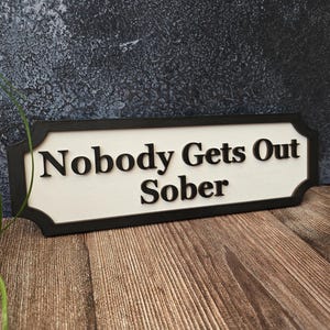May include: A rectangular sign with the text "Nobody Gets Out Sober" in black, set against a white background. The sign has a black border and is placed on a wooden surface.
