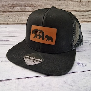 May include: Black trucker hat with a brown leather patch that says "PAPA" with a bear and cub silhouette. The hat has a black mesh back.
