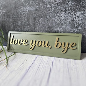 May include: A rectangular olive green sign featuring the raised gold-coloured text "love you, bye" in a cursive script. The sign has a clean, flat border and is positioned horizontally, displaying a smooth finish on the lettering and the base.