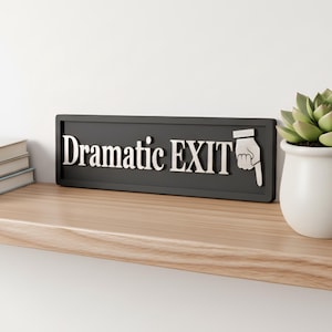 May include: A rectangular black sign with raised silver lettering that reads "Dramatic EXIT" followed by a silver icon of a hand with a pointing index finger. The sign is positioned horizontally on a wooden surface next to a small green succulent.