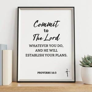 May include: A rectangular black frame containing a white print with the black text: Commit to The Lord WHATEVER YOU DO, AND HE WILL ESTABLISH YOUR PLANS. PROVERBS 16:3, featuring a small black cross symbol next to the scripture reference.