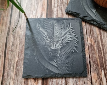 Puede incluir: Un posavasos de pizarra negra con una imagen grabada detallada de una cabeza de dragón. La boca del dragón está abierta, revelando dientes afilados. El grabado es de color gris, creando un efecto 3D.