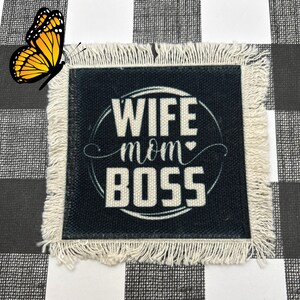 May include: Square black coaster with white fringe and the words "WIFE MOM BOSS" in white. A yellow and black butterfly is in the upper left corner. The coaster is on a black and white checkered surface.