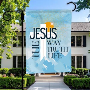 May include: A garden flag with the words "JESUS THE WAY TRUTH LIFE" in white and black text, with a white cross and a yellow sunburst design. The flag is displayed in front of a white house with black shutters.