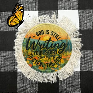 May include: A round, white, fringed coaster with a colorful sunset scene and the text "God is still writing your story. Stop trying to steal the pen."