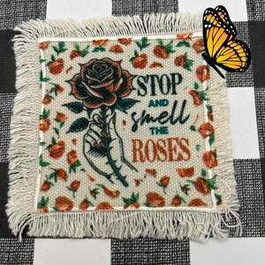 May include: A white fabric patch with frayed edges features a hand holding a red rose with green leaves. The text "STOP AND SMELL THE ROSES" is printed in black and brown lettering.