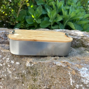 May include: A rectangular stainless steel lunch box with a light brown bamboo lid. The lid is secured with black metal clasps. The lunch box is sitting on a large, textured rock, with green foliage in the background.