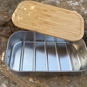 May include: A rectangular, stainless steel lunch box with a bamboo lid. The lid is light brown and has rounded edges. The lunch box is open, revealing a silver interior with vertical ridges. The lunch box is sitting on a rock.