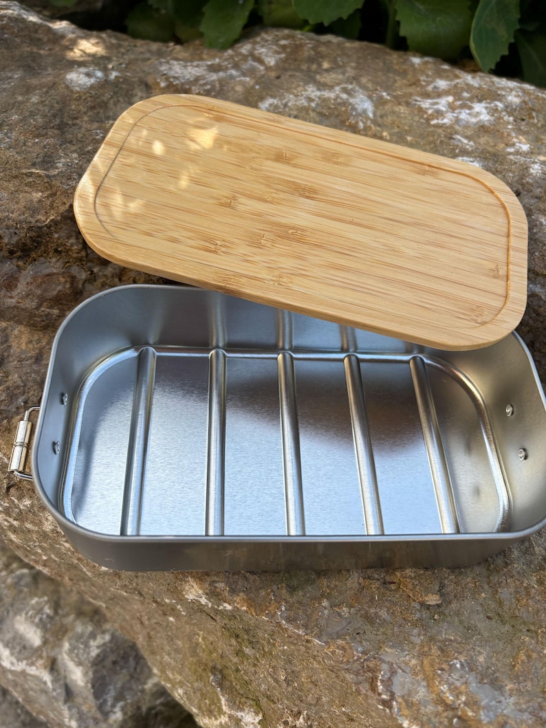 May include: A rectangular, stainless steel lunch box with a bamboo lid. The lid is light brown and has rounded edges. The lunch box is open, revealing a silver interior with vertical ridges. The lunch box is sitting on a rock.