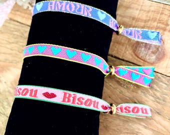 Op de afbeelding: Drie kleurrijke vriendschapsarmbanden met tekst- en hartmotieven. De bovenste armband is blauw met het woord "AMOUR" in roze. De middelste armband is roze met turquoise hartjes. De onderste armband is roze met het woord "Bisou" en een rood lipdesign.